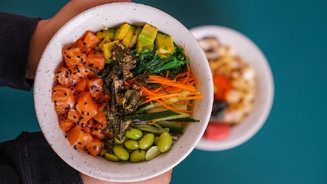Sepasang tangan memegang semangkuk poke bowl dengan salmon dan potongan sayur
